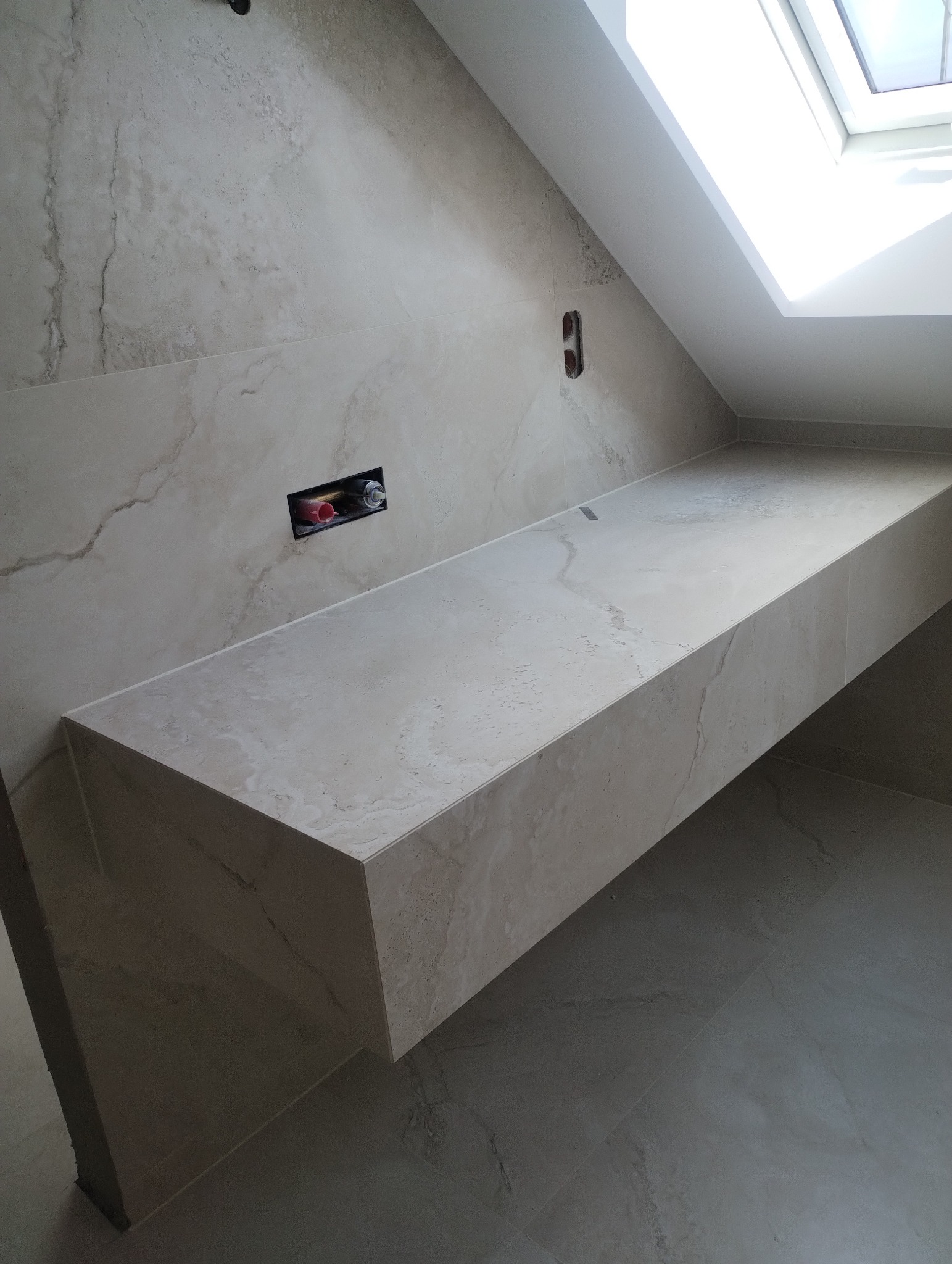 Beżowa łazienka na poddaszu ze skośnym oknem. Wykończona zlewem z płytek imitujących marmur, z widocznymi przyłączami wodnymi i odpływem liniowym.