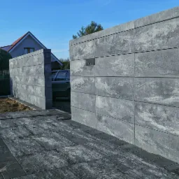 Nowoczesna brama wjazdowa z szarej kostki betonowej, z widocznym fragmentem posesji i kostką brukową na podjeździe, w tle dom z czerwonym dachem.