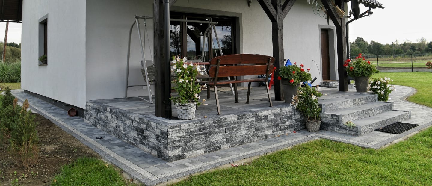 Wykończony taras z kostki brukowej w Nysie, z niskim murem z dekoracyjnej cegły, schodami i ścieżką, otoczony zielonym trawnikiem i kwitnącymi kwiatami w donicach.