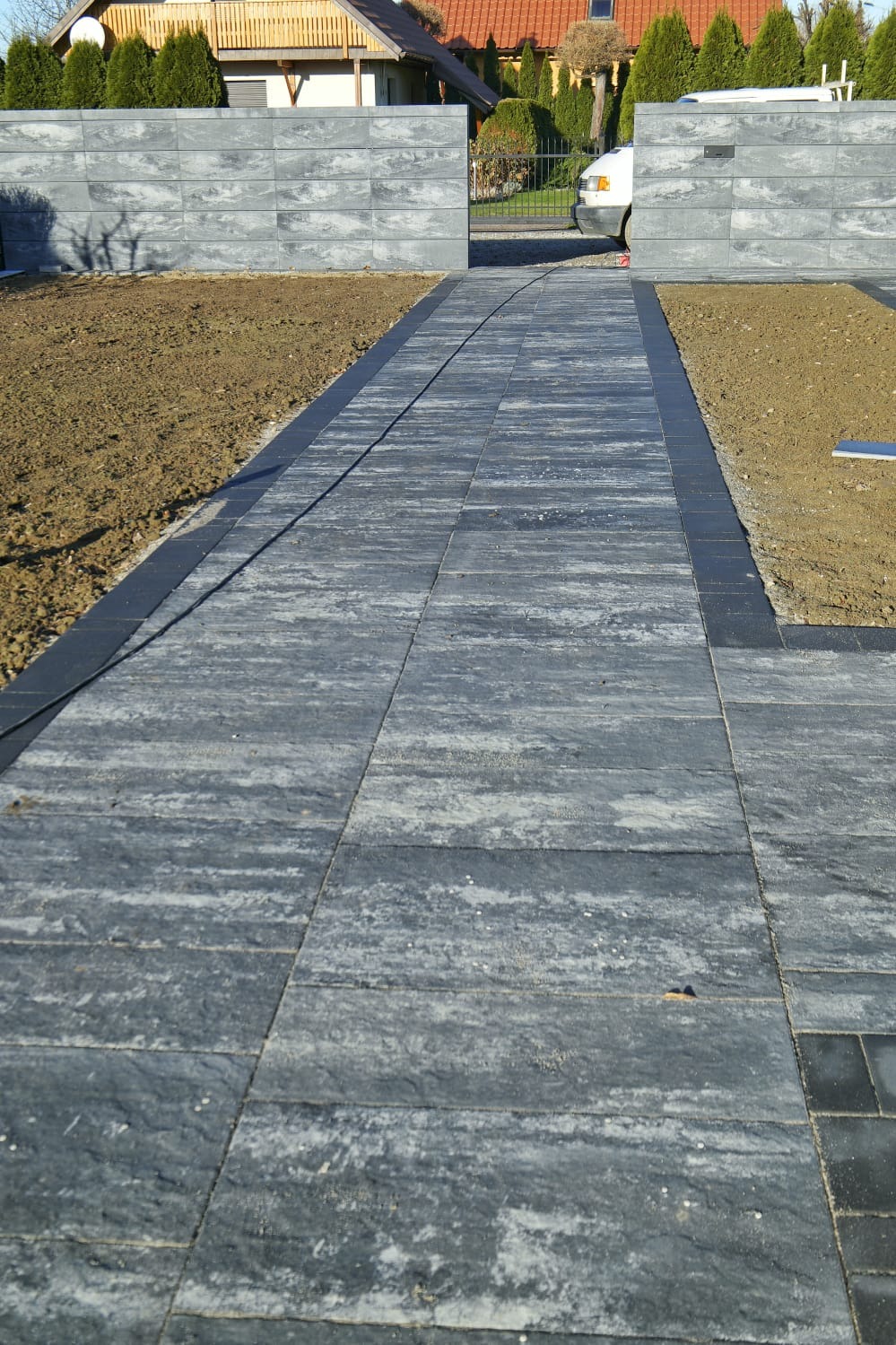 Ułożona, szara kostka brukowa tworząca ścieżkę wjazdową, obrzeża z ciemnej kostki, po bokach przygotowany teren pod trawnik, w tle brama wjazdowa i dom.