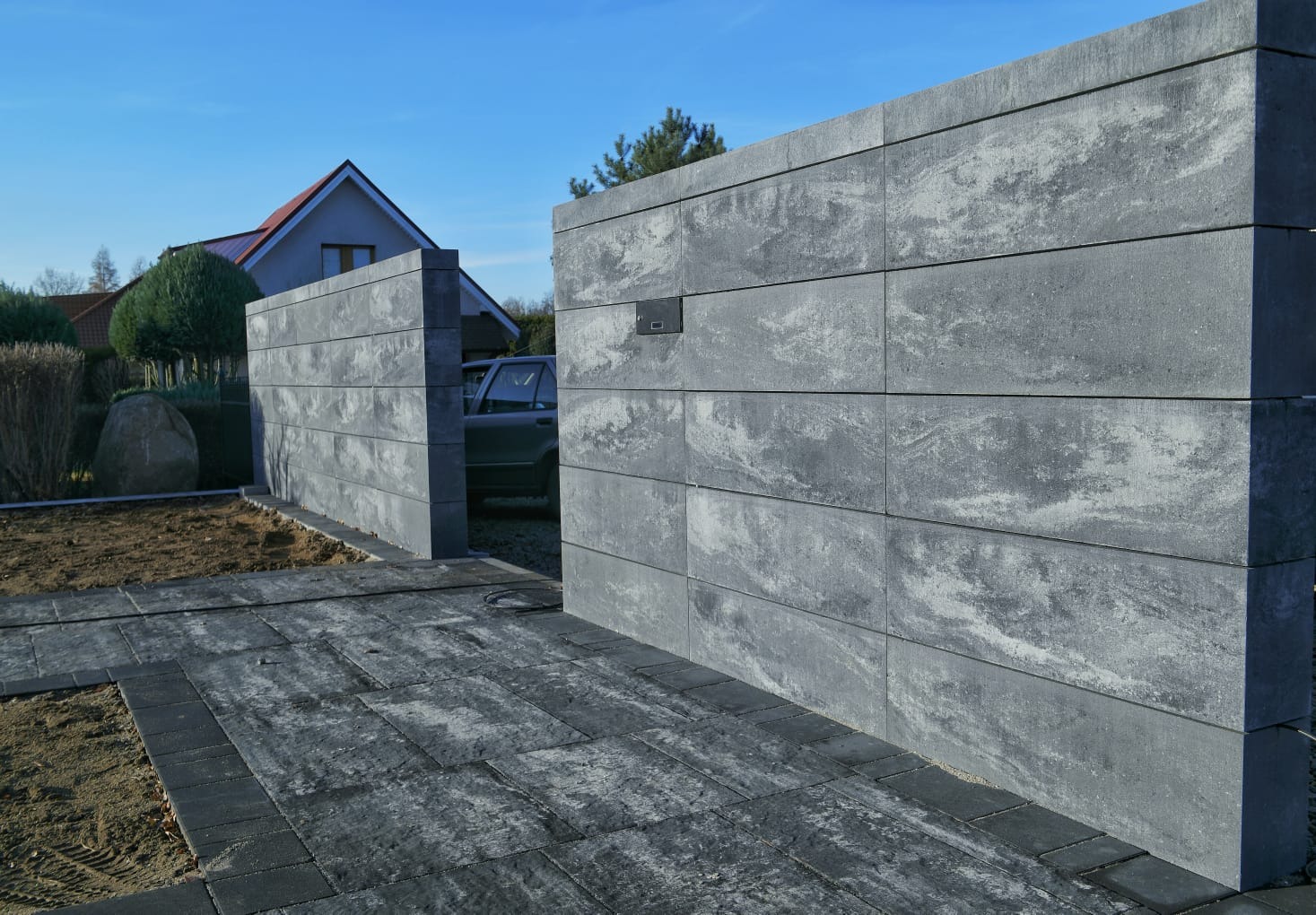 Nowoczesna brama wjazdowa z szarej kostki betonowej, z widocznym fragmentem posesji i kostką brukową na podjeździe, w tle dom z czerwonym dachem.