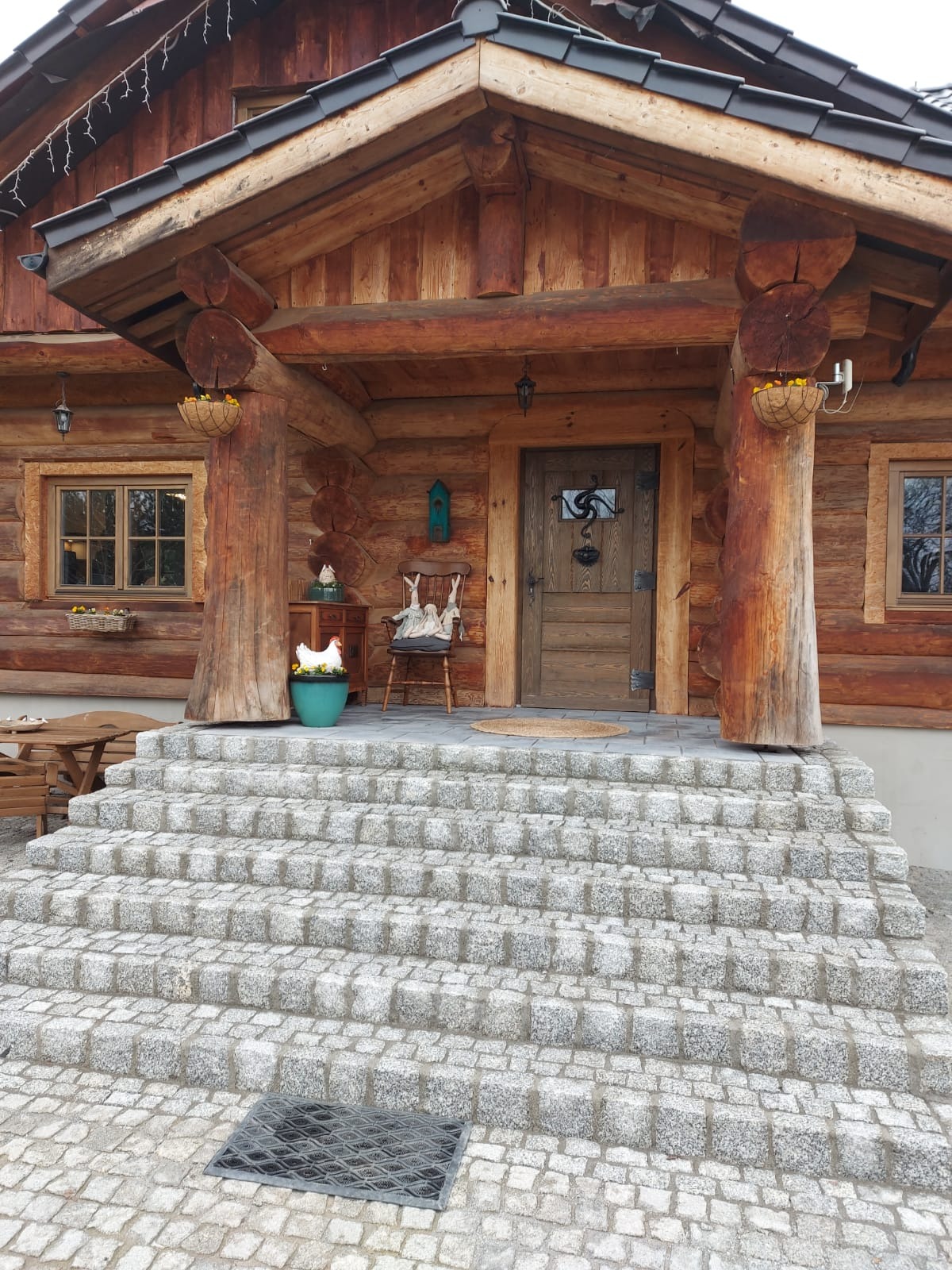 Schody wejściowe do drewnianego domu wyłożone kostką granitową, widok na front domu z drewnianymi elementami i dekoracjami.