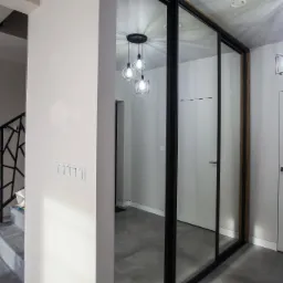 Nowoczesne wnętrze: szklana ściana działowa z czarną ramą, odbijająca wiszące lampy w stylu industrialnym, obok schody z geometryczną balustradą i szarym dywanem.