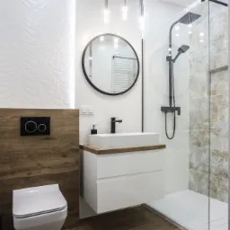 Nowoczesna łazienka z kabiną prysznicową, umywalką nablatową na szafce z szufladami i okrągłym lustrem w czarnej ramie, akcenty drewniane i białe płytki z tłoczeniami.