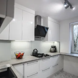 Biała, nowoczesna zabudowa kuchenna z szarym blatem, czarnym zlewem i płytą gazową, oświetleniem LED pod szafkami wiszącymi i koszem z owocami na blacie.