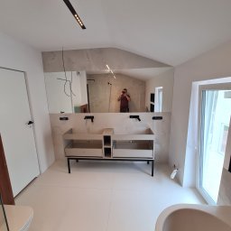 Firma remontowo-wykonczeniowa Hubert Lach - Nowoczesna łazienka w trakcie wykańczania: podwójna umywalka na czarnym stelażu, duże lustro z odbiciem fotografa, białe płytki podłogowe, częściowo widoczna wanna i toaleta, minimalistyczne...