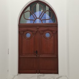 Firma remontowo-wykonczeniowa Hubert Lach - Eleganckie, dwuskrzydłowe, drewniane drzwi wewnętrzne z łukowym przeszkleniem w kształcie kwiatu, osadzone w białej ścianie nad marmurową podłogą.