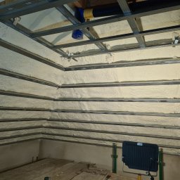 Firma remontowo-wykonczeniowa Hubert Lach - Poddasze ocieplone pianą PUR pomiędzy metalowymi profilami konstrukcyjnymi, widoczna lampa halogenowa i fragment podłogi z desek.