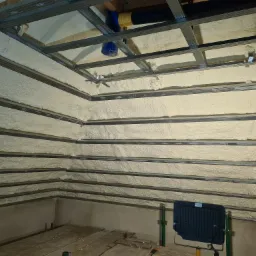 Poddasze ocieplone pianą PUR pomiędzy metalowymi profilami konstrukcyjnymi, widoczna lampa halogenowa i fragment podłogi z desek.