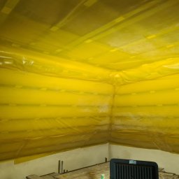 Firma remontowo-wykonczeniowa Hubert Lach - Poddasze zabezpieczone żółtą folią przed pracami wykończeniowymi, widoczne łączenia folii taśmą, fragment podłogi z deskami i grzejnik elektryczny.