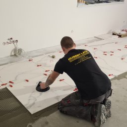 Firma remontowo-wykonczeniowa Hubert Lach - Glazurnik w czarnej koszulce z logo firmy Hubert Lach układa białe płytki imitujące marmur na podłodze, używając przyssawki do płytek i czerwonych krzyżyków dystansowych.