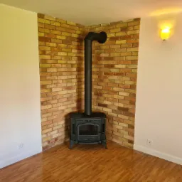 Czarny piec wolnostojący z rurą odprowadzającą spaliny, umieszczony w rogu pokoju wyłożonym cegłą, na podłodze panele imitujące drewno, ściany pomalowane na jasny kolor.