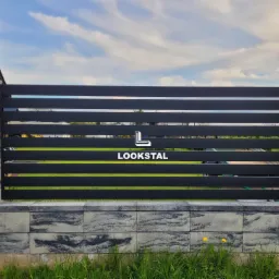 Nowoczesne ogrodzenie z poziomych, czarnych profili, osadzone na słupkach z szarej kostki imitującej kamień, z widocznym logo firmy 'LOOKSTAL' na tle błękitnego nieba.