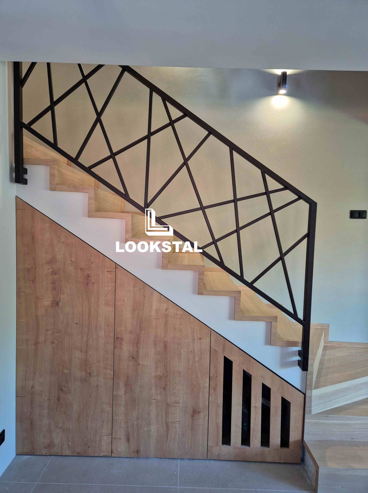 Nowoczesna poręcz schodowa z geometrycznym wzorem, wykonana z ciemnego metalu. Schody drewniane z zabudową szafki pod schodami. Oświetlenie punktowe na ścianie.