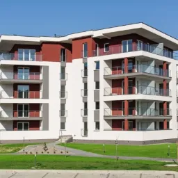 Nowoczesny budynek wielorodzinny z balkonami o szklanych balustradach, pomalowany na biało z bordowymi akcentami, widoczny z perspektywy trawnika i parkingu w słoneczny dzień.