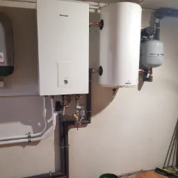 Biały kocioł gazowy Panasonic Aquarea z podłączonymi rurami i zbiornikiem Galmet, zamontowany na ścianie w kotłowni; widoczne również naczynie wzbiorcze i fragment innego kotła po lewej stronie.