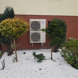 Biała jednostka zewnętrzna klimatyzacji Panasonic Inverter zamontowana na ceglanej ścianie budynku, otoczona ozdobnymi drzewami i białym żwirem.