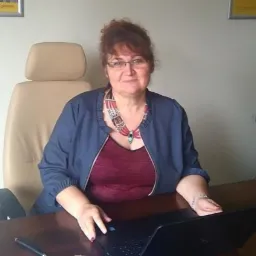 Kobieta w okularach i naszyjniku siedzi za biurkiem z laptopem w biurze, w tle widoczny plakat.