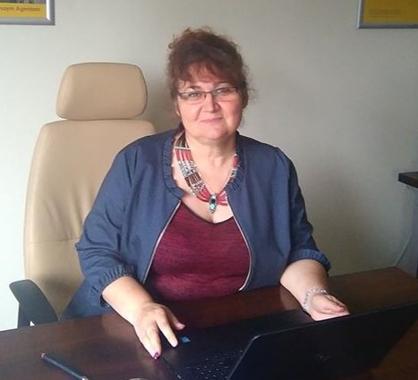 Kobieta w okularach i naszyjniku siedzi za biurkiem z laptopem w biurze, w tle widoczny plakat.