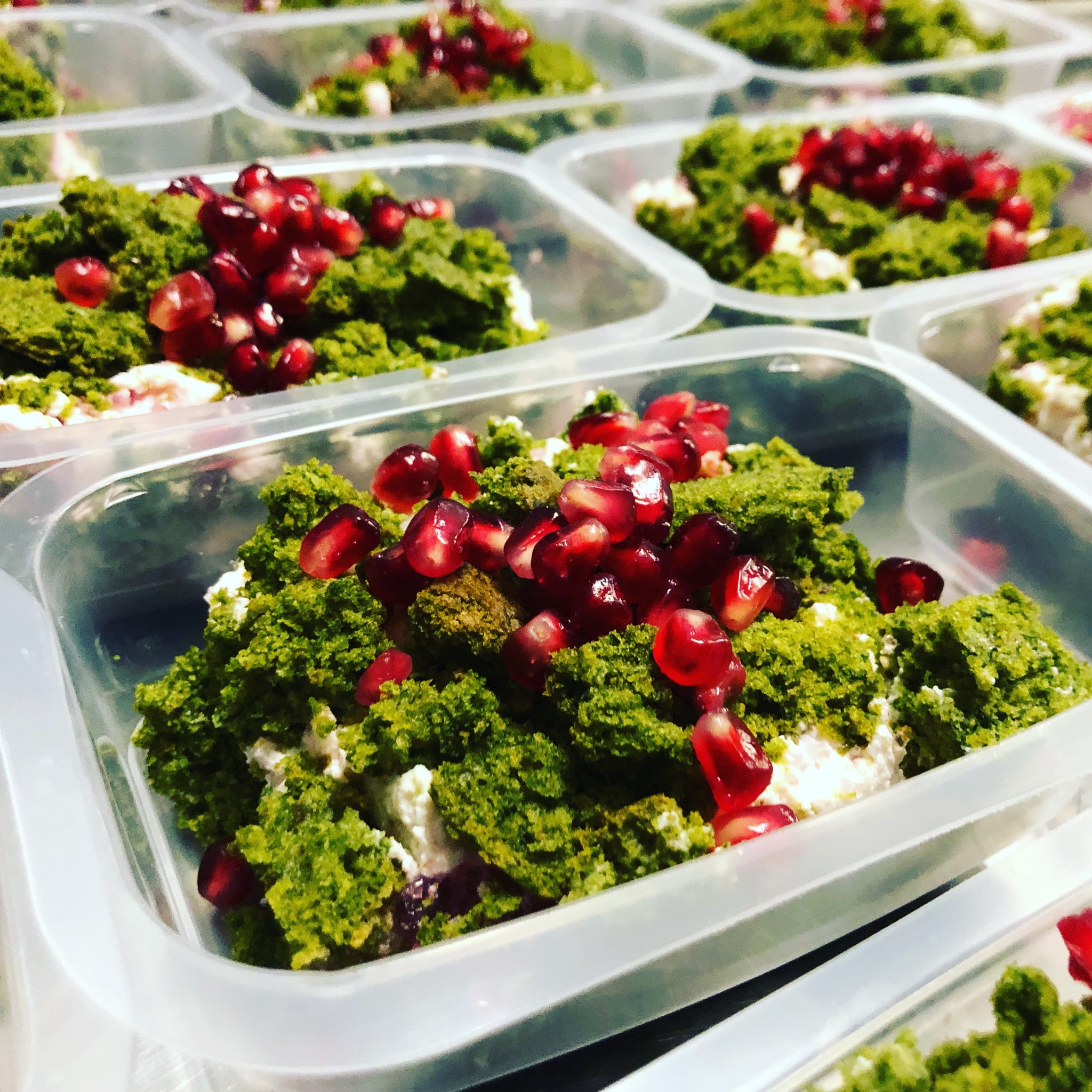 Porcje sałatki z zielonych warzyw, sera feta i granatu w przezroczystych pojemnikach na wynos, ułożone rzędami na metalowym blacie.