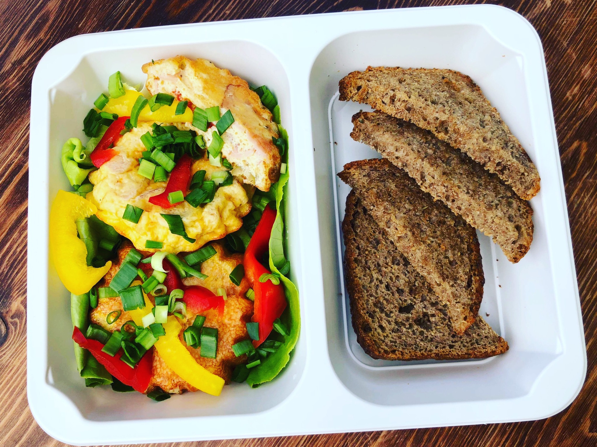 Dietetyczny posiłek w białym pojemniku: omlet warzywny z papryką i szczypiorkiem oraz trzy kromki ciemnego pieczywa na drewnianym stole.