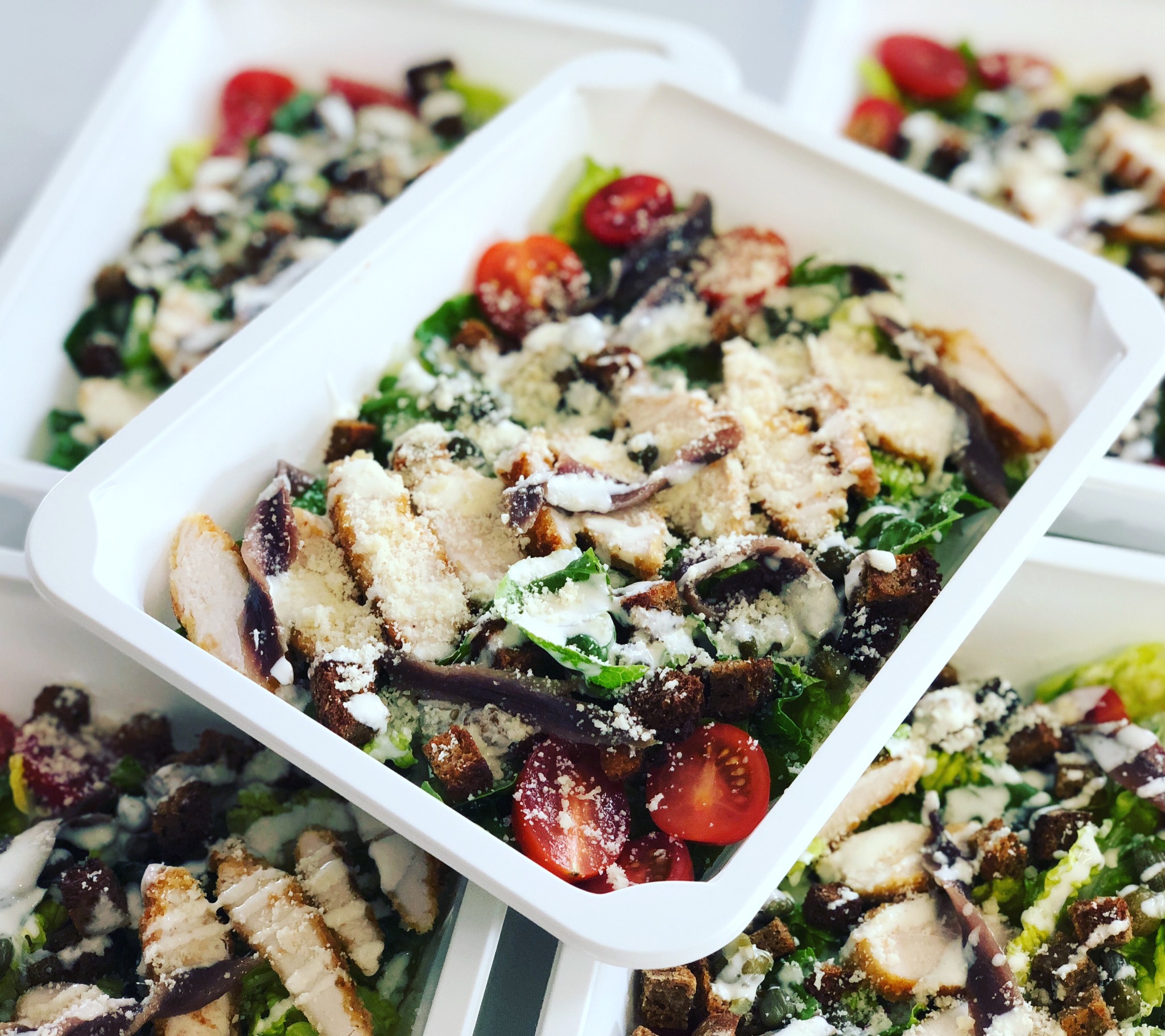 Sałatka Cezar z grillowanym kurczakiem, pomidorkami cherry, grzankami, anchois i parmezanem, podana w białym, plastikowym pojemniku.