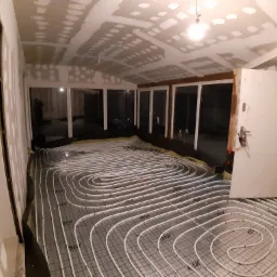 Rozłożone rury ogrzewania podłogowego na folii izolacyjnej, widoczne ściany w trakcie wykańczania płytami kartonowo-gipsowymi, surowy stan.