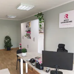 Wnętrze biura rachunkowego: biurko z monitorem AOC, krzesło biurowe, tablica z logo 'Biuro Rachunkowe z pasji do księgowości', biała szafa z książkami i roślinami, doniczkowe kwiaty na biurku...