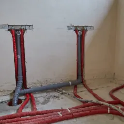 Surowy stan instalacji: szare rury kanalizacyjne i czerwone rury grzewcze w ścianie, z widocznymi metalowymi uchwytami mocującymi i fragmentami gruzu na podłodze.