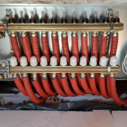 Rozdzielacz ogrzewania podłogowego z czerwonymi rurami i białymi zaworami, widoczne metalowe elementy montażowe oraz zwój białych kabli z boku.