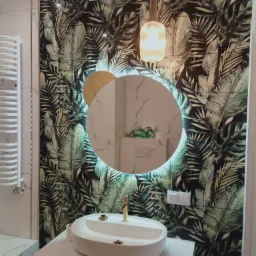 Nowoczesna łazienka z okrągłym lustrem podświetlanym LED i płytkami w tropikalne liście, złota armatura, biała umywalka nablatowa i grzejnik drabinkowy.