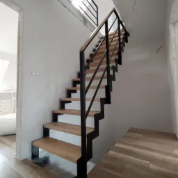 Nowoczesne schody z drewnianymi stopniami i czarną metalową konstrukcją, widoczne w jasnym wnętrzu domu w trakcie wykończenia, z widocznymi elementami instalacji elektrycznej na suficie i logo...