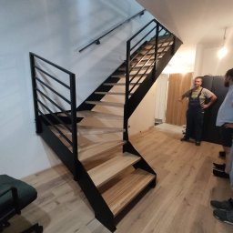 bertaweld - Nowoczesne schody metalowe z drewnianymi stopniami w jasnym wnętrzu, wykonane na zamówienie, widoczni pracownicy w tle.