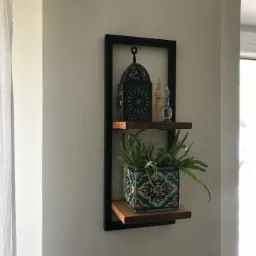 Półka wisząca z czarną ramą, drewnianymi blatami, na których stoi dekoracyjny lampion, flakoniki i doniczka z rośliną o długich liściach, całość na tle białej ściany.