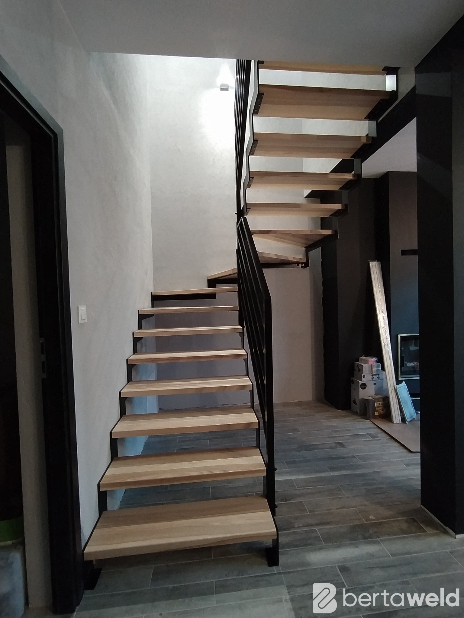 Nowoczesne schody z drewnianymi stopniami i czarną metalową balustradą, widok z dołu na jasne, wysokie pomieszczenie z szarą ścianą i ciemnymi elementami wystroju.