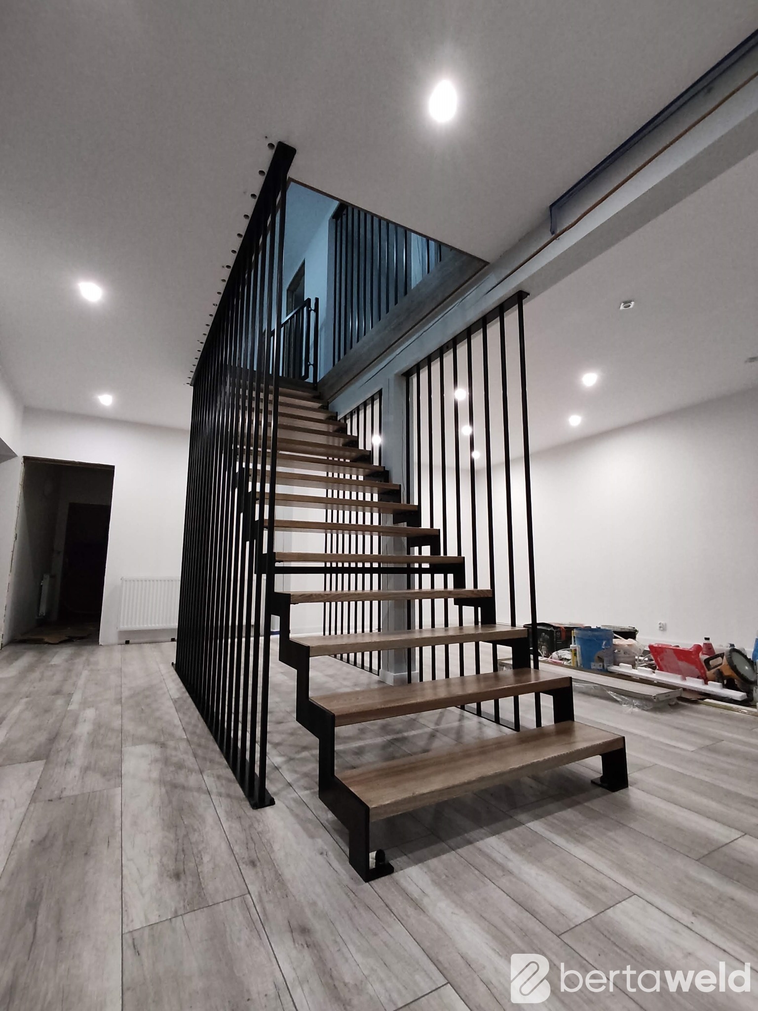 Nowoczesne schody z drewnianymi stopniami i czarną metalową balustradą w jasnym wnętrzu, widoczne narzędzia i materiały budowlane na podłodze.