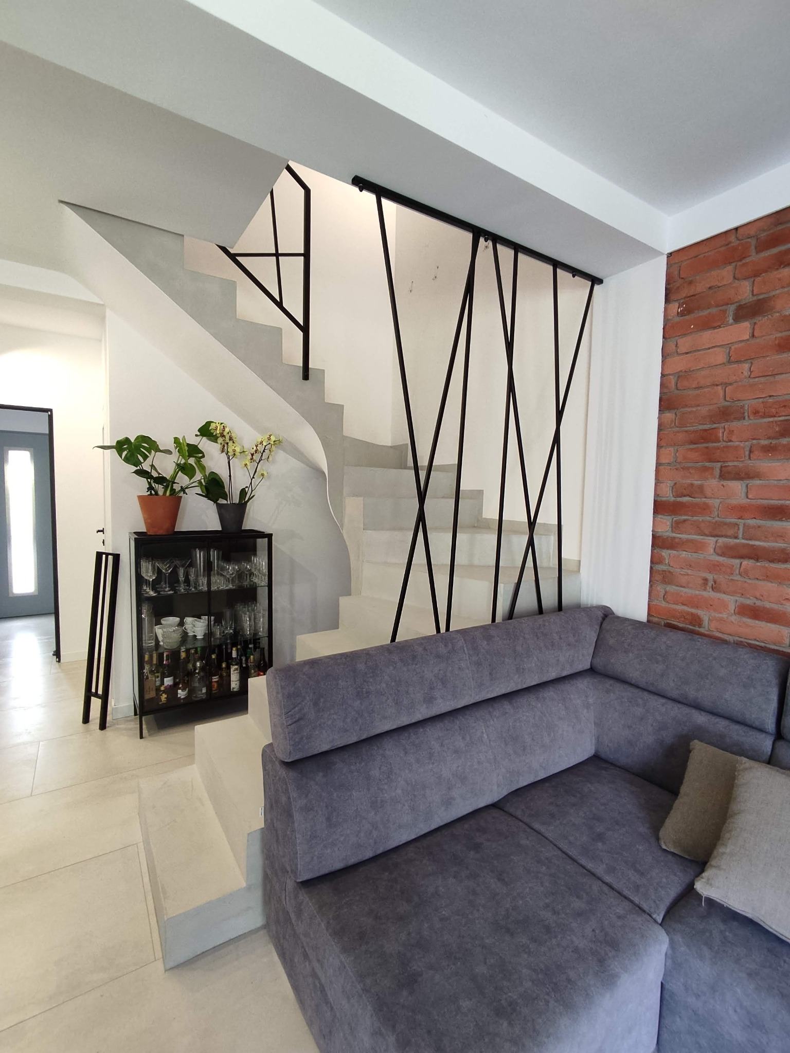 Betonowe schody z czarną, geometryczną balustradą w nowoczesnym wnętrzu z sofą i ścianą z czerwonej cegły.