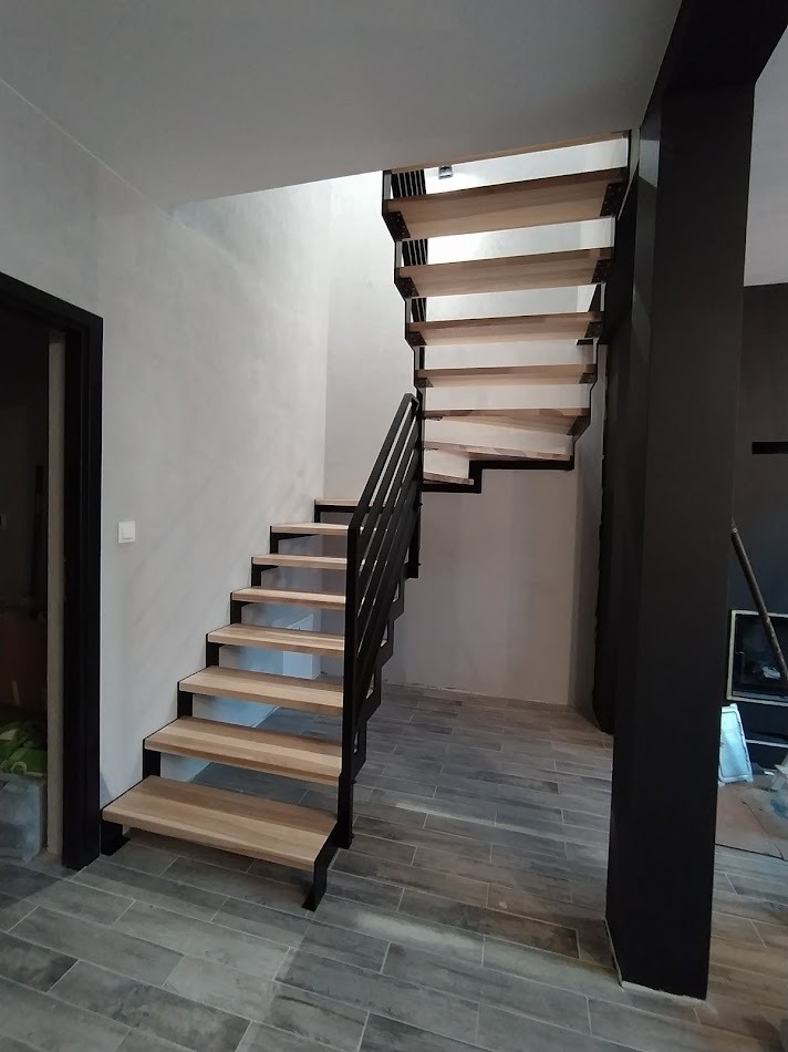Schody metalowe z drewnianymi stopniami, czarne metalowe barierki, szare płytki podłogowe, minimalistyczny design, nowoczesne wnętrze w trakcie wykańczania.