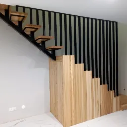 Nowoczesne schody z drewnianymi stopniami i czarną, pionową balustradą, zintegrowane z zabudową pod schodami z forniru o jasnym usłojeniu, na tle jasnych ścian i marmurowej podłogi.