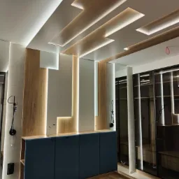 Nowoczesna sypialnia z drewnianym sufitem podwieszanym z oświetleniem LED, zabudową meblową z tapicerowanym wezgłowiem w kolorze granatowym i szklaną szafą wnękową.