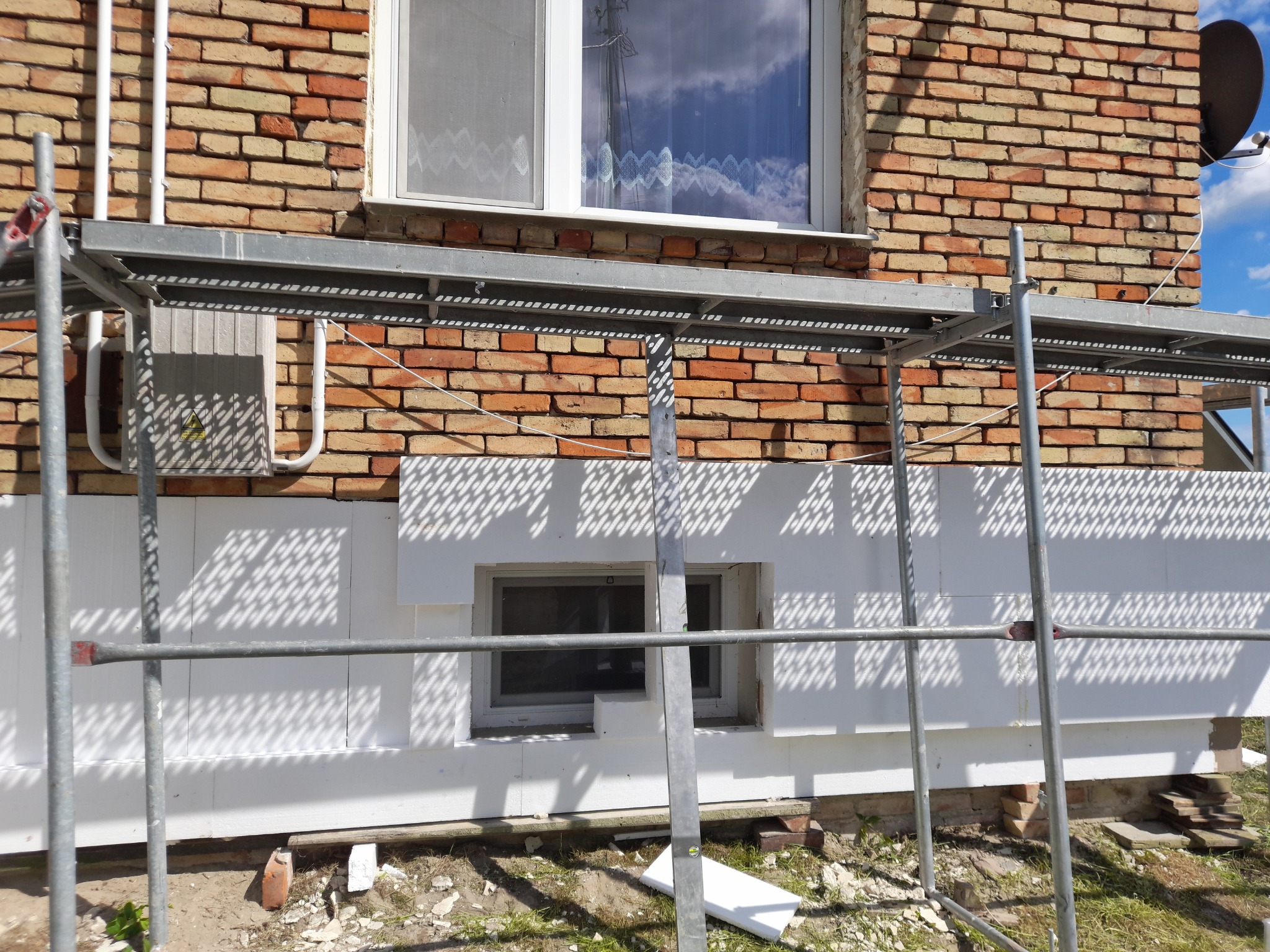 Dom z cegły z zamontowanym rusztowaniem i częściowo ocieploną elewacją styropianem, widoczne okno z odbijającymi się chmurami na szybie.