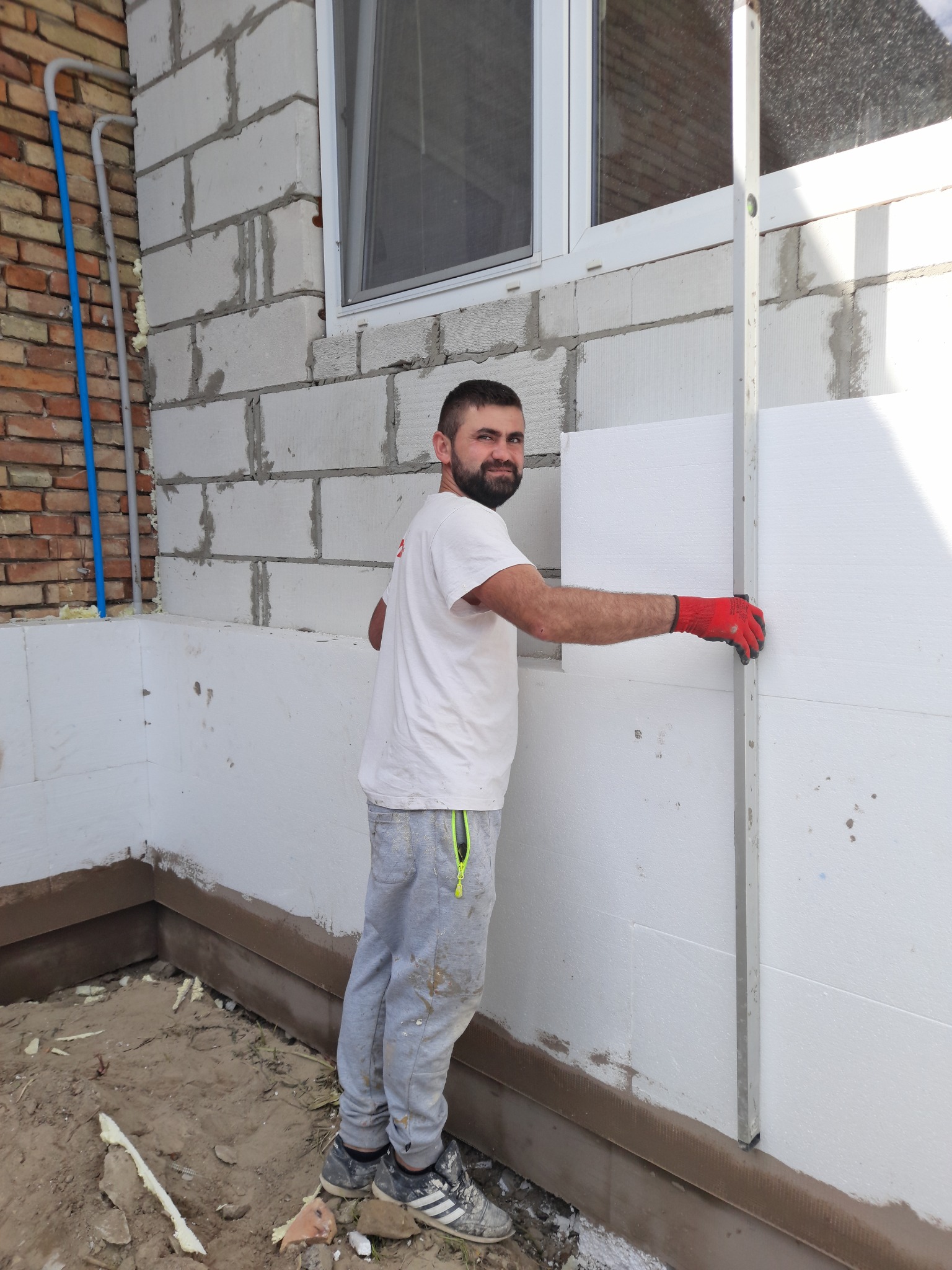 Mężczyzna w czerwonych rękawicach poziomuje płytę styropianową na ścianie budynku z białego bloczka, widoczny fragment muru z cegły i okno.