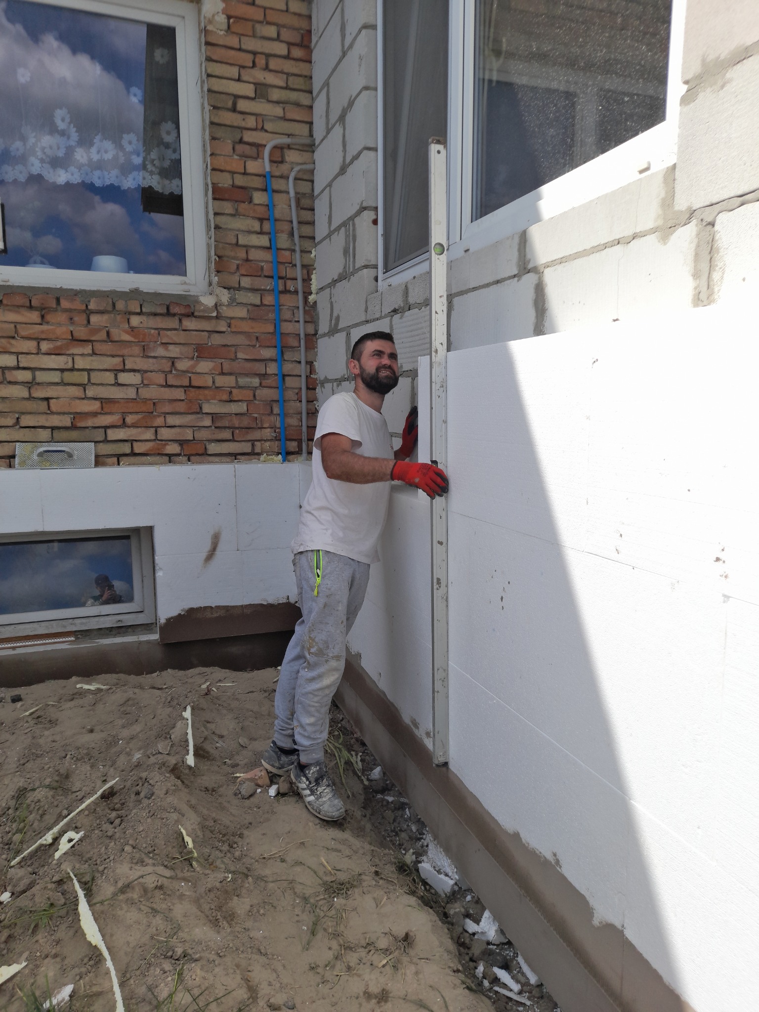 Mężczyzna w czerwonych rękawicach poziomuje białe płyty styropianowe na ścianie budynku z cegły i bloczków, widoczne okno z odbiciem nieba i ziemia z resztkami styropianu u podstawy.