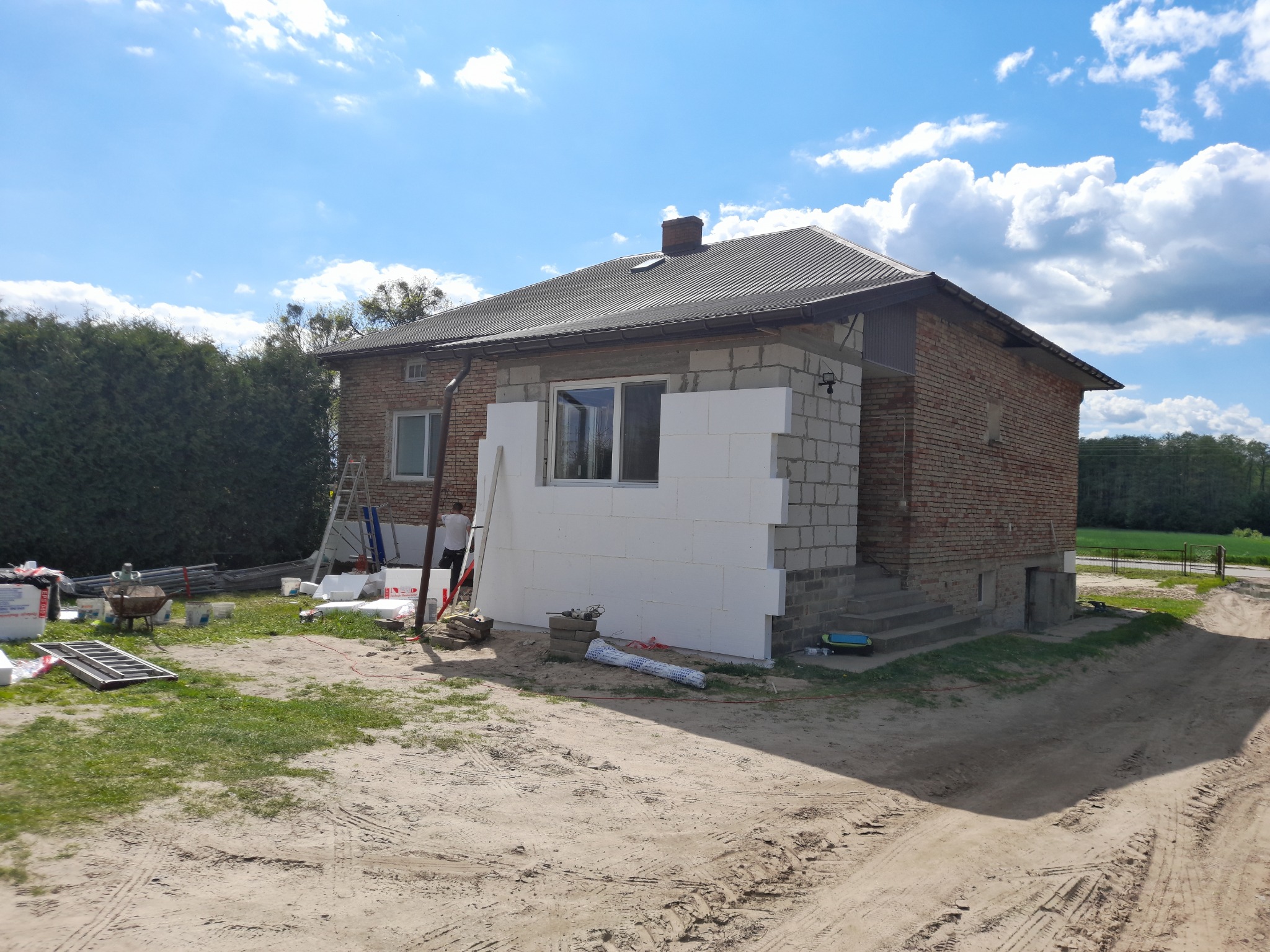 Dom z cegły w trakcie ocieplania, widoczne białe płyty styropianowe na ścianie, rusztowania i narzędzia budowlane w otoczeniu, słoneczny dzień.