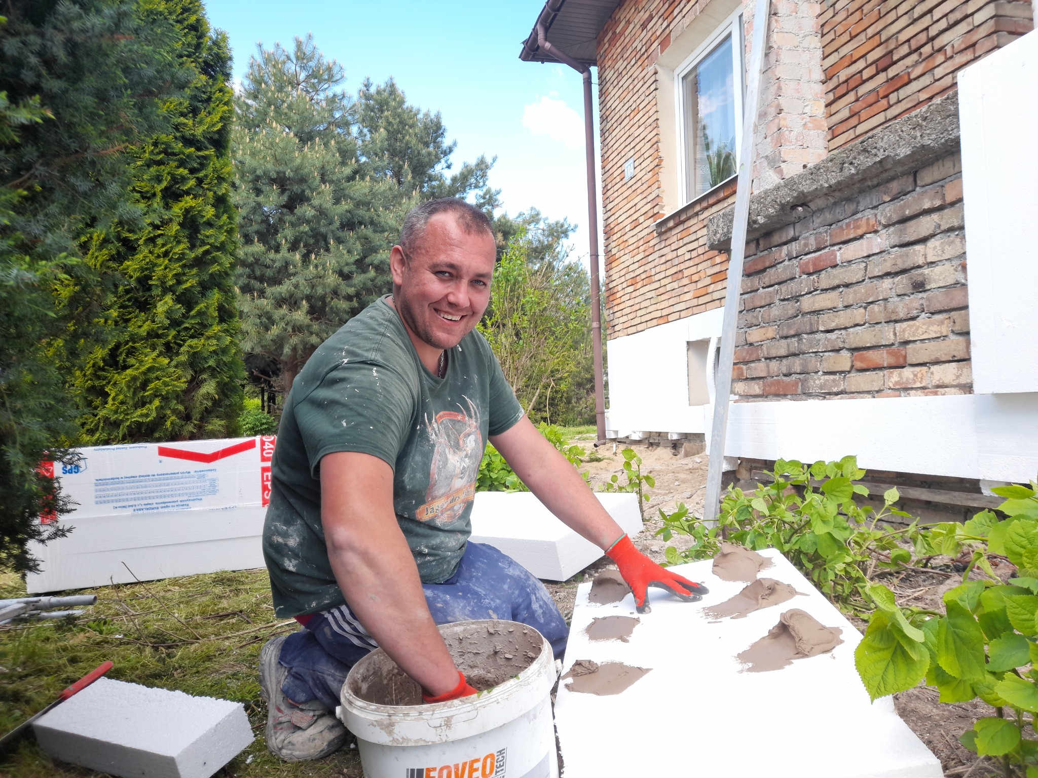 Uśmiechnięty mężczyzna w czerwonych rękawicach nakłada zaprawę na styropianową płytę, przygotowując ją do ocieplenia elewacji domu z cegły.