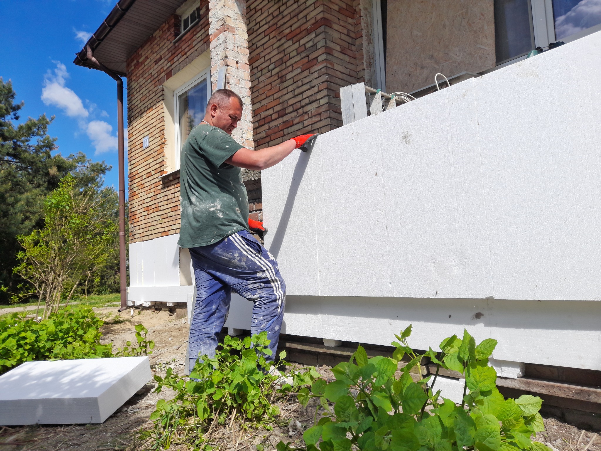 Pracownik w czerwonych rękawicach montuje białe płyty styropianowe na elewacji domu z cegły, widoczne rusztowanie i zieleń.