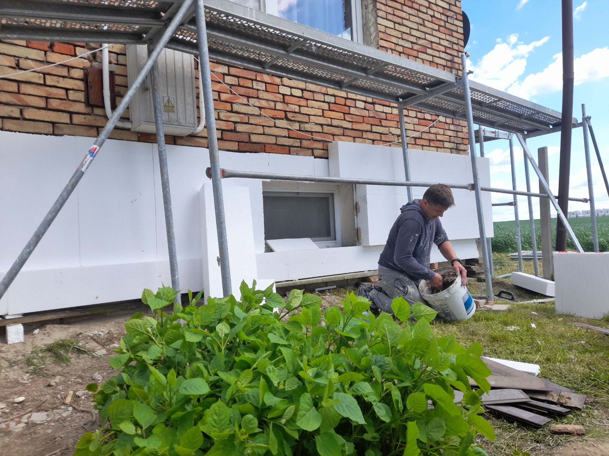 Pracownik na kolanach przygotowuje zaprawę pod rusztowaniem przy ocieplonej styropianem ścianie budynku z czerwonej cegły, widoczny fragment elewacji i bujna zieleń.