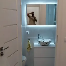 Nowoczesna łazienka z białą szafką pod umywalkę, umywalką w stylu lastryko, podświetlanym lustrem i heksagonalnymi płytkami podłogowymi; w lustrze odbija się osoba robiąca zdjęcie.