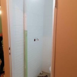 Adam Kurzawa - Wnętrze łazienki w trakcie remontu, widoczne świeżo położone białe płytki, narzędzia na podłodze i wystające kable. Drzwi wejściowe częściowo zasłaniają widok.