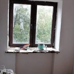 Adam Kurzawa - Pokój w trakcie odświeżania: widoczne okno z brązową ramą, przygotowane narzędzia malarskie na parapecie, surowe ściany przed malowaniem i prowizoryczne oświetlenie.
